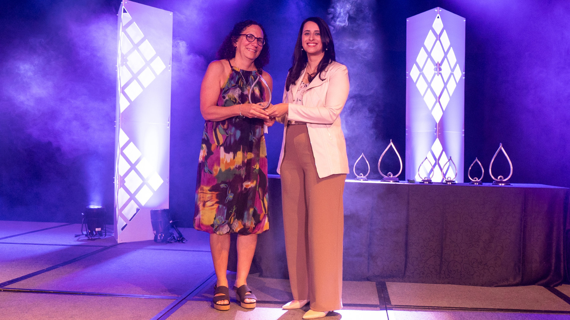 Two women stand on a stage, with their hands together. One woman is handing the other an award. 