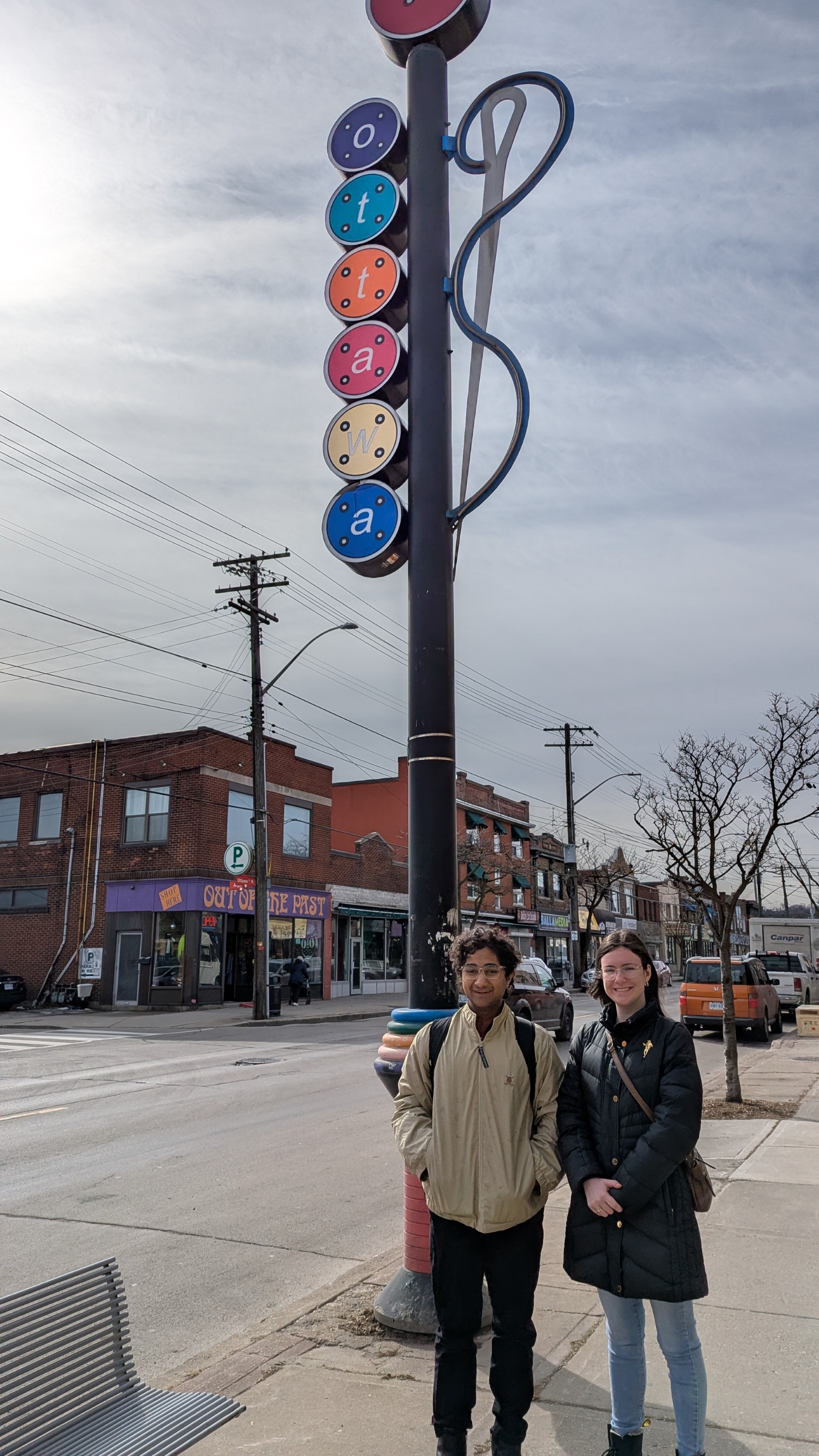 Two young people, wearing coats, stand on a sidewalk of a city street. Low red brick buildings are visible across the street behind them. Above their heads, a signpost reads 'Ottawa.' 