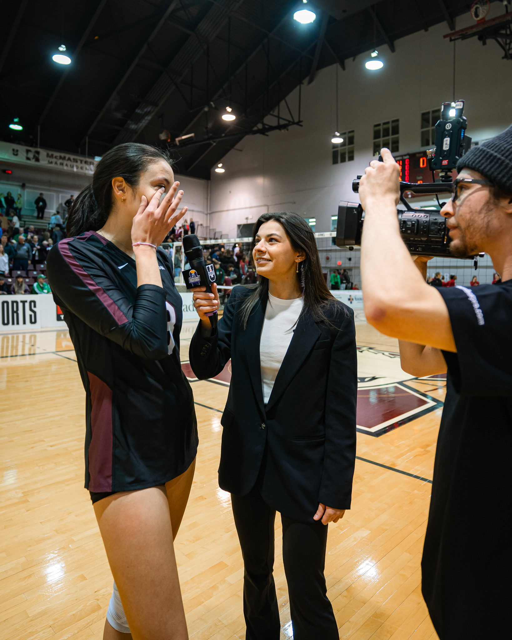 A woman with a microphone interviews an emotional volleyball player on the side of a court. A cameraman is filming them. 