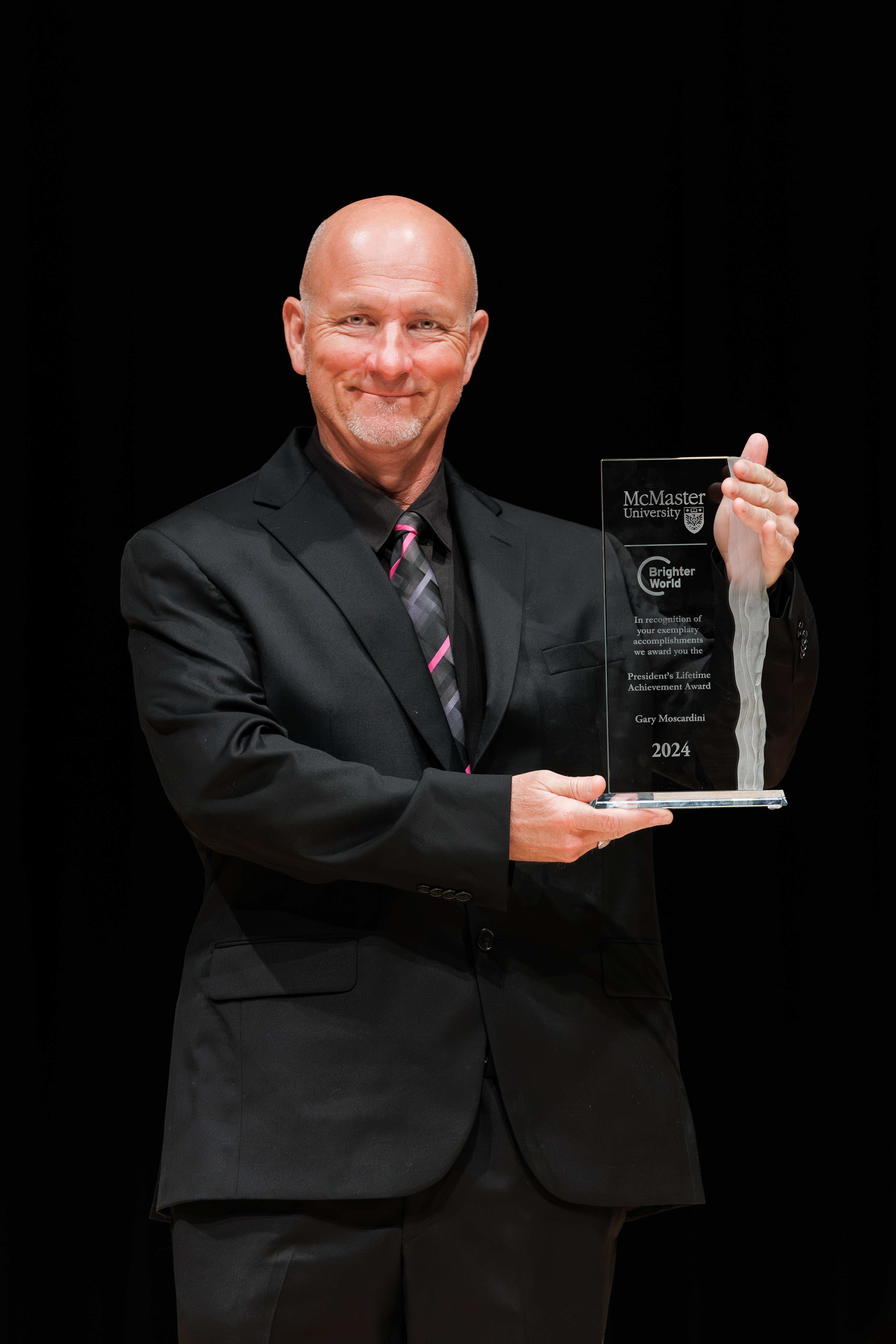 Gary Moscardini, a smiling man in a dark suit and a tie, holds up his glass award against a dark backdrop.