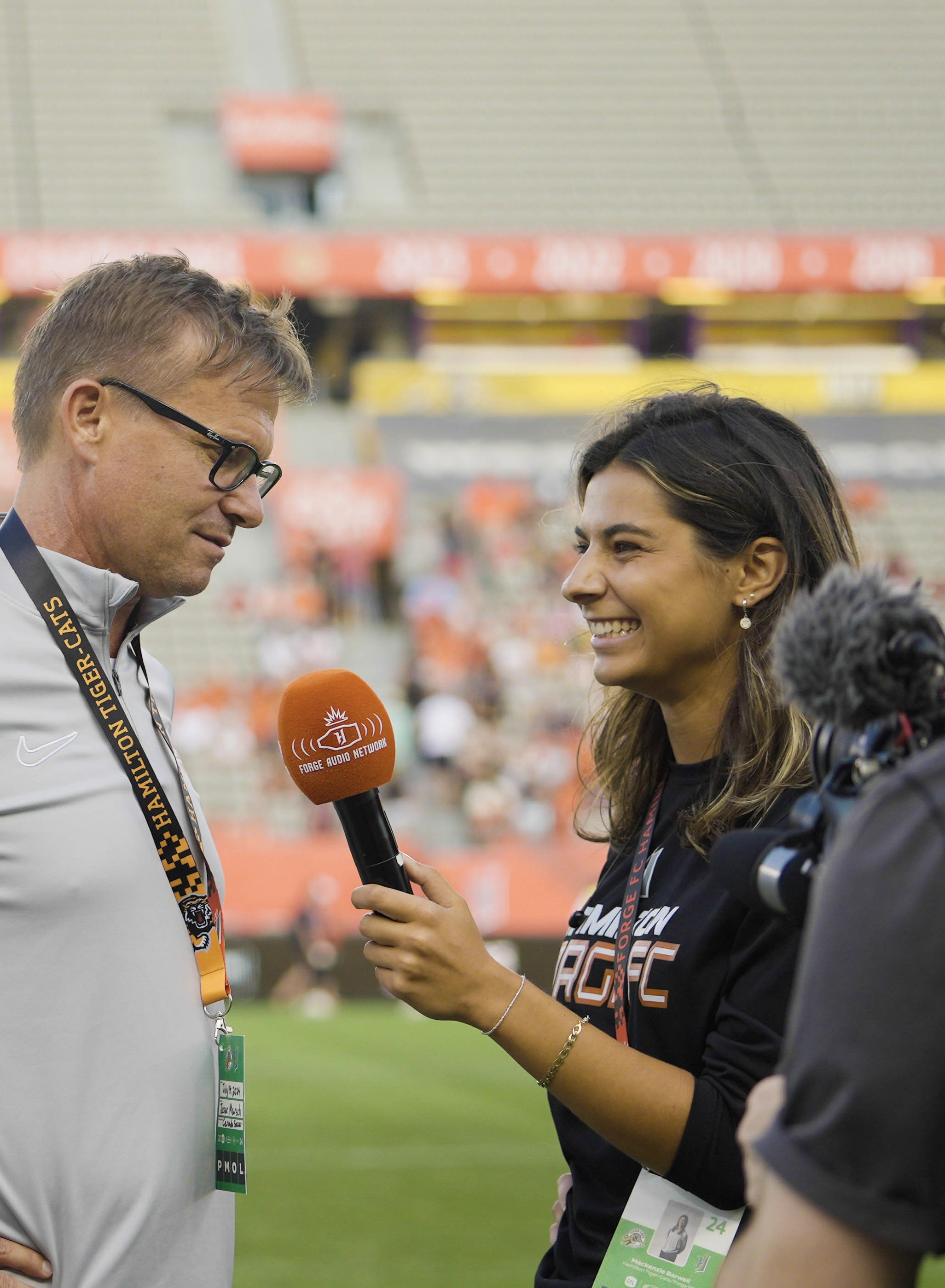 A woman with a microphone interviews someone on the edge of a sporting field.