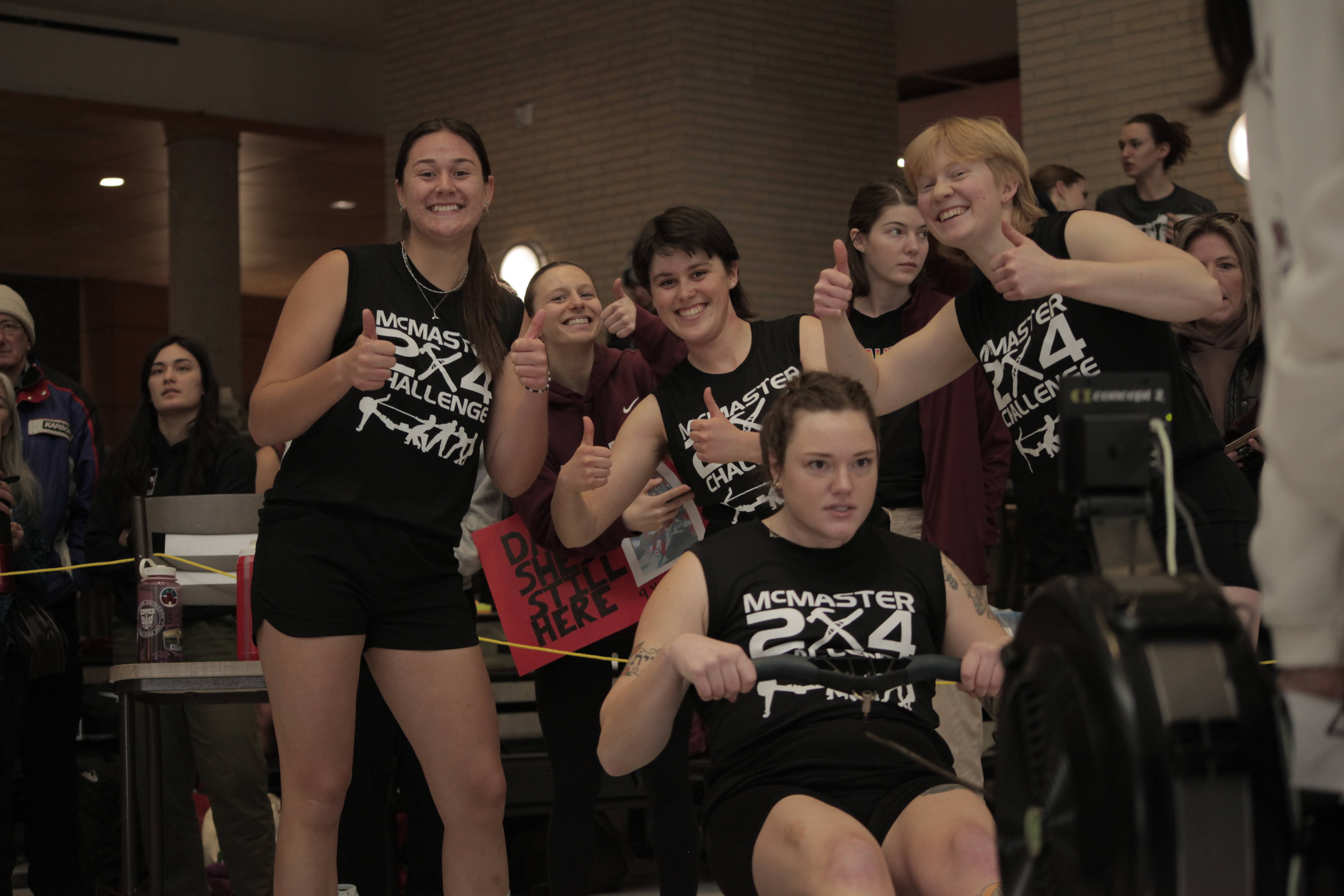 Six women gather around a rowing machine, with one of them seated on the machine.