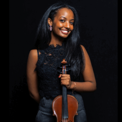 A woman in dress clothes holds a violin. The background behind her is black. 