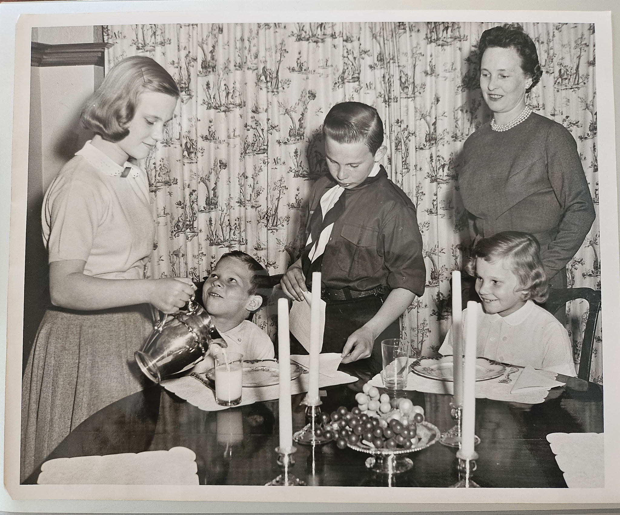 A black and white photo of a woman and four children at a dinner table, laid with food and candles.