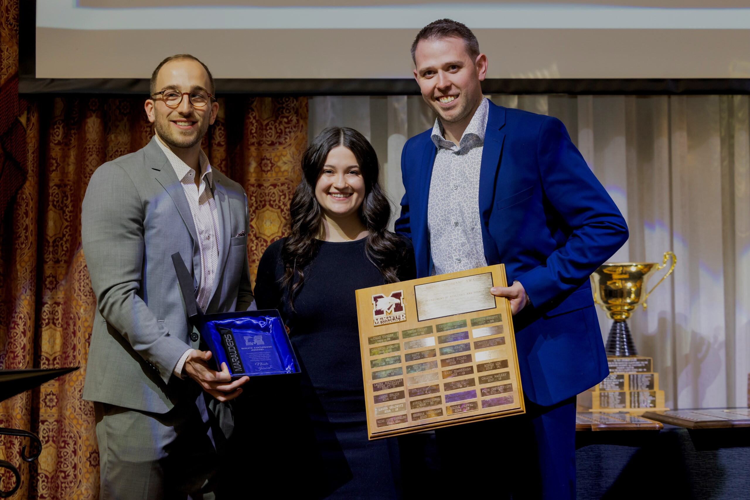 A woman and two men in suits stand together, holding an award and award plaque.