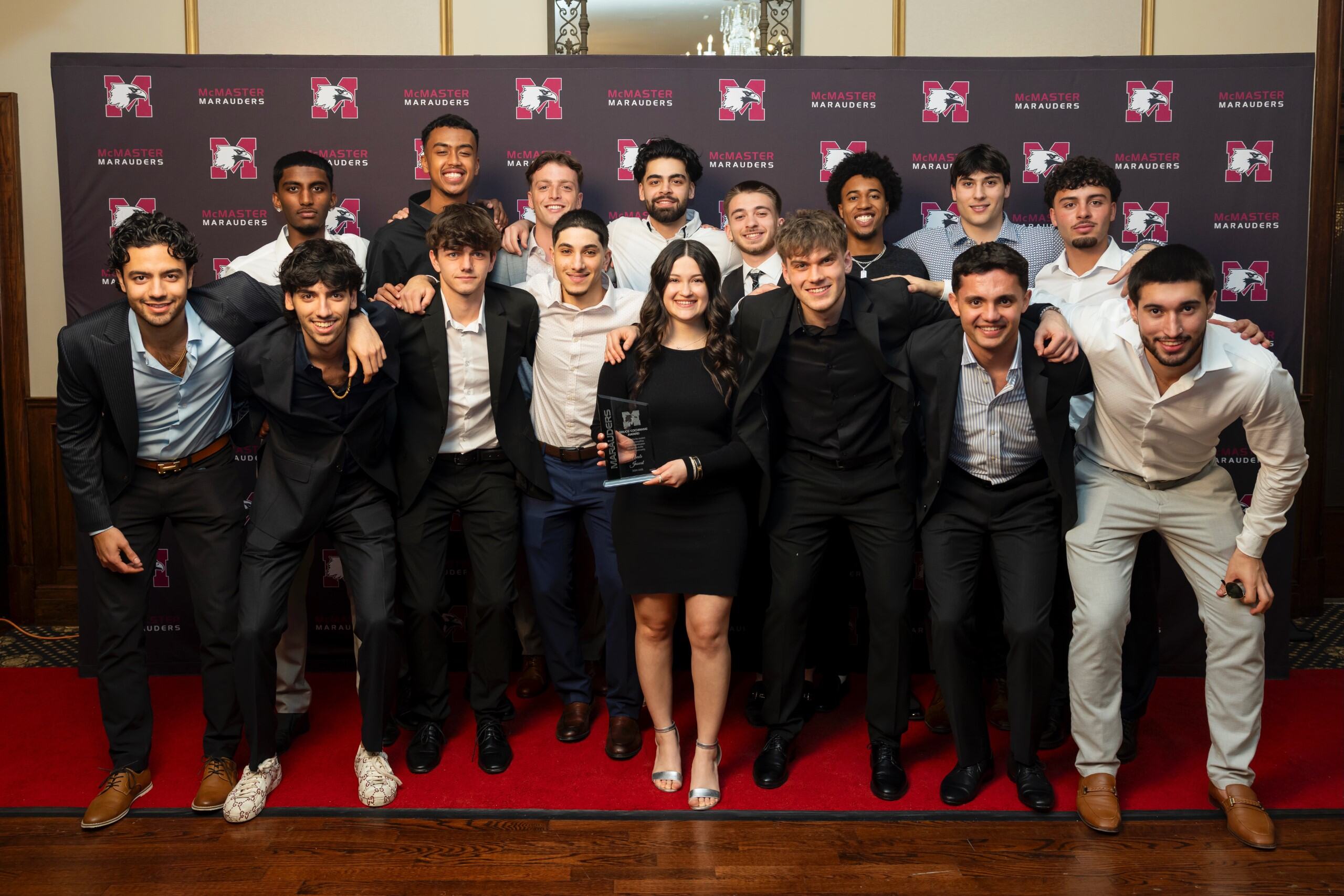 A woman holding a trophy and a group of men, posing for a picture in front of a red backdrop.