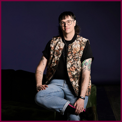 A headshot of a person sitting on the arm of a green couch. They are wearing jeans, a black t-shirt, glasses, and a colourful vest.