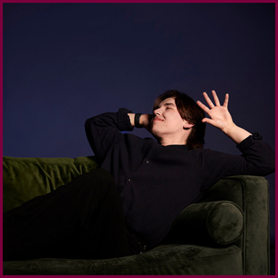 A headshot of a person lying on a green couch, one hand near their head with their palm facing up and outwards.
