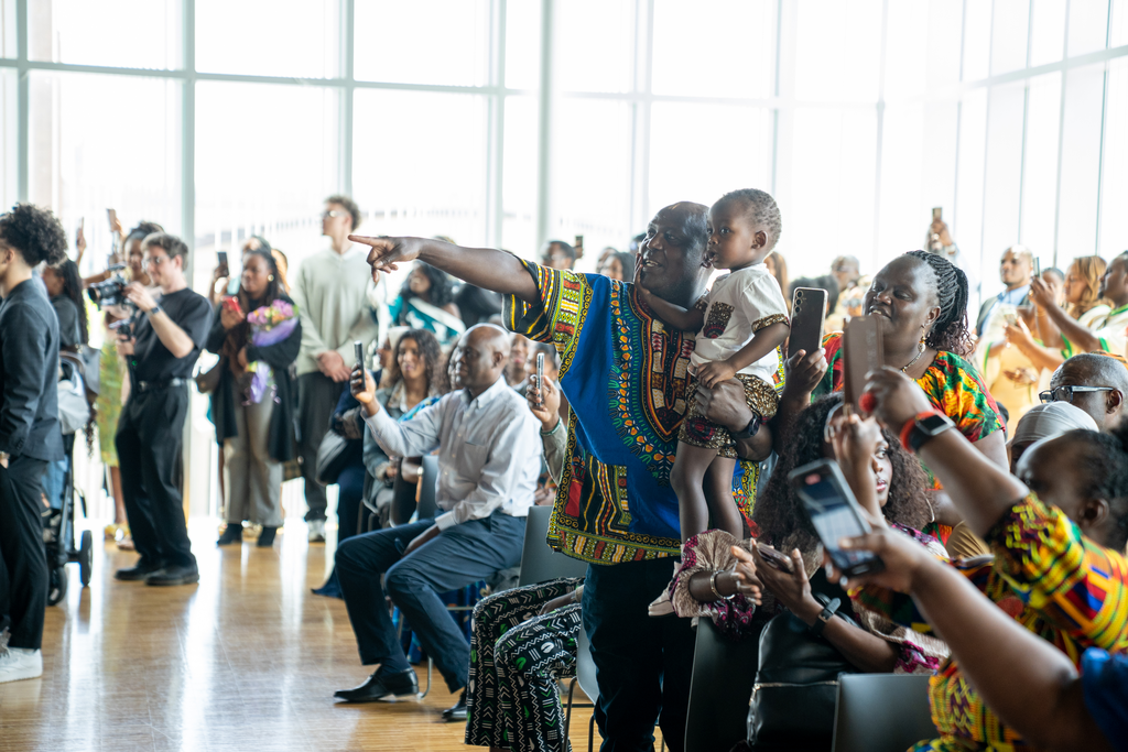 A man holds a child, pointing to something. They are in a crowd of people, many of them holding up phones to take photos.