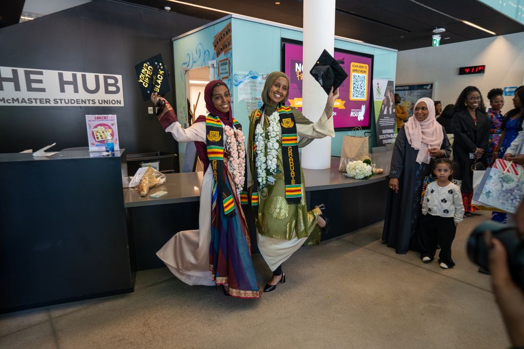 Two women in dresses pose, each holding up a small black signboard with gold letters. 