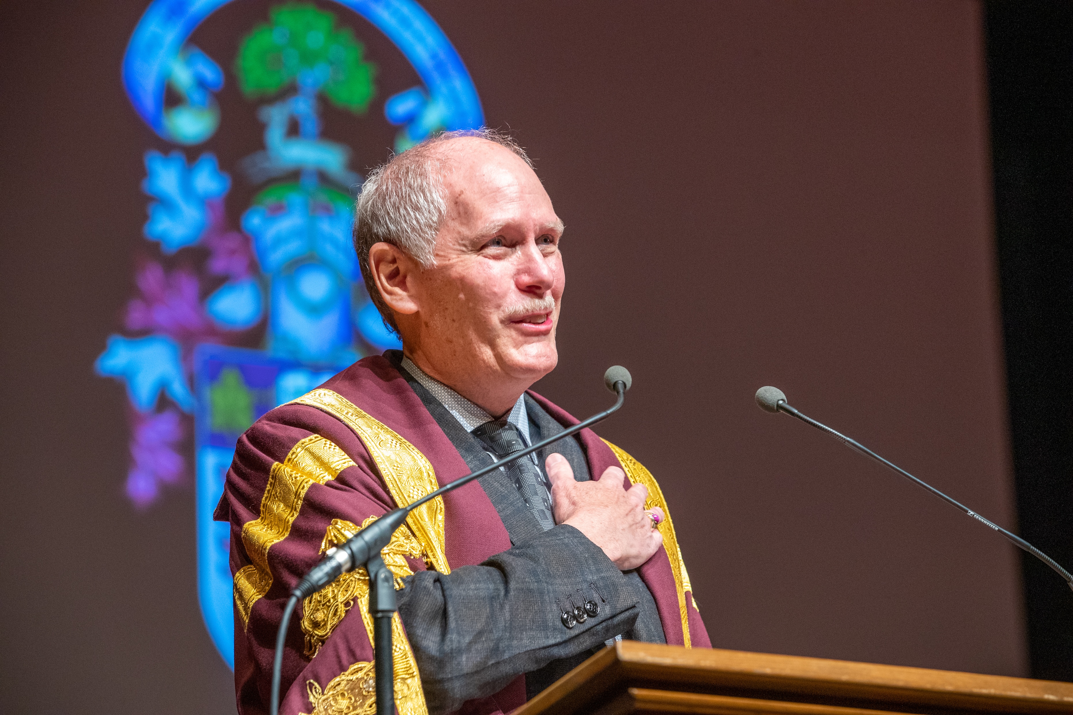 David Farrar, in McMaster University President's ceremonial robe, places his hand on his heart and smiles as he speaks to graduating students on stage at convocation.