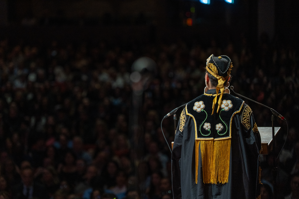 The back of a woman dressed in robes, embroided with flowers and golden thread. She is standing at a podium, facing a crowd in a theatre. 