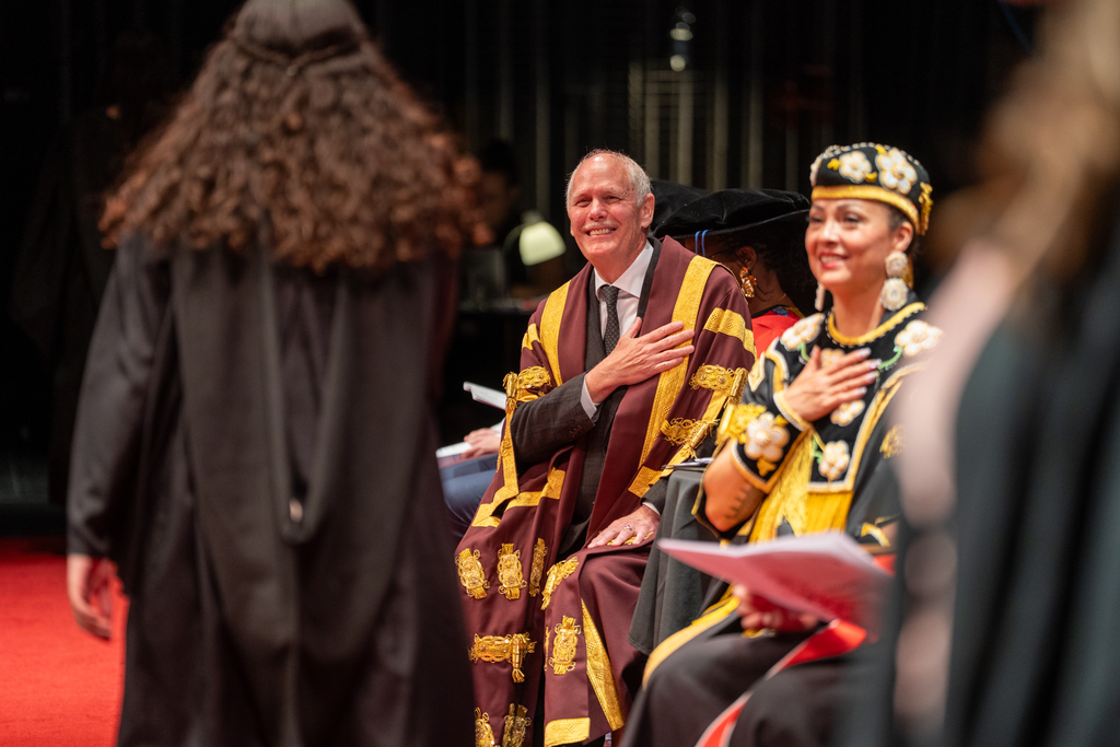 Two people in robes each hold a hand over their heart, watching a person in robes walk in front of them.