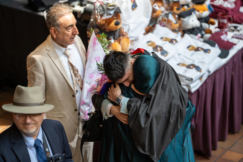A man in graduation robes and a woman embrace. Another man stands just behind them, holding flowers.