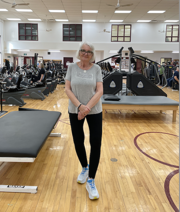 A grey-haired woman with glasses, wearing a tshirt, leggings and sneakers in a room full of fitness equipment.
