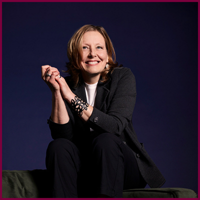 A headshot of a person sitting on a green couch and smiling widely.