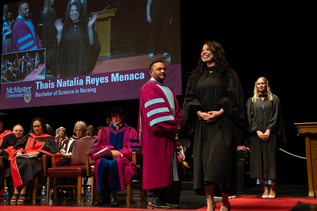 A woman in graduation robes beams, standing on a stage.