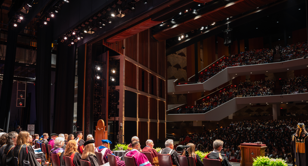 An auditorium, with about twenty people sitting on a stage, looking out over a packed audience sitting across three levels. 
