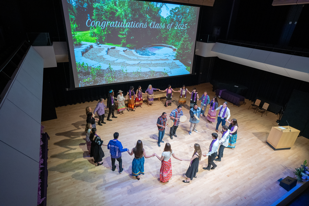 A group of people dance in a circle on a stage, holding hands. In the centre of the circle are three men with drums. A large screen above them reads 'Congratulations Class of 2025.'