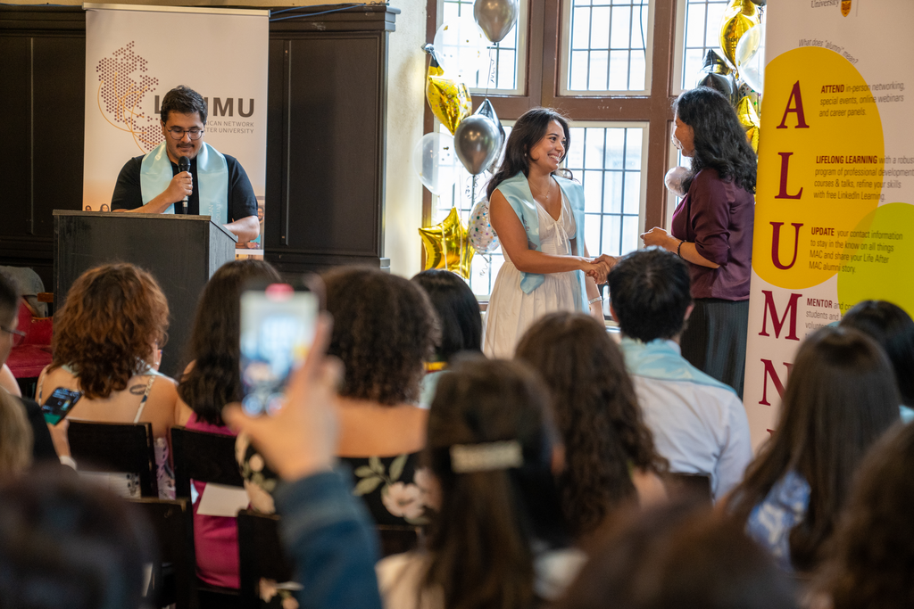 Amidst a crowd of people, somebody holds their phone up to take a photo of two people, who are standing at the front of the room.