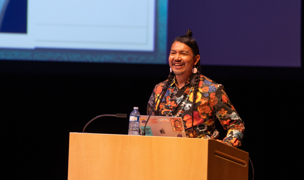 A person stands at a lectern, with a presentation screen behind him.