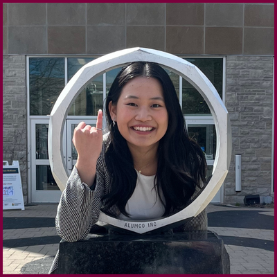 A headshot of a woman posing by a metal ring, holding up her smallest finger on her right hand.