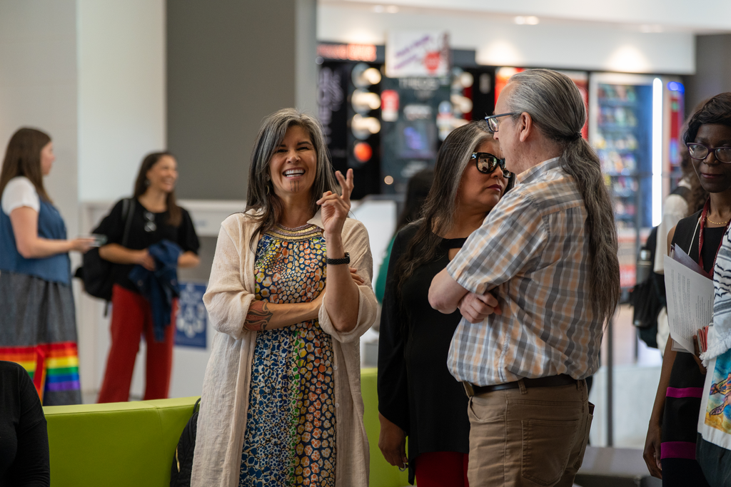 Three people stand in a semi-circle in an indoor space, with more people visible behind them. One woman is smiling at the camera.