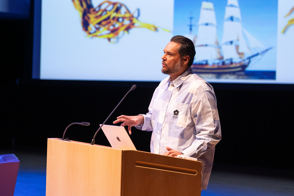 A person stands at a lectern, with a presentation screen showing a large ship behind him.
