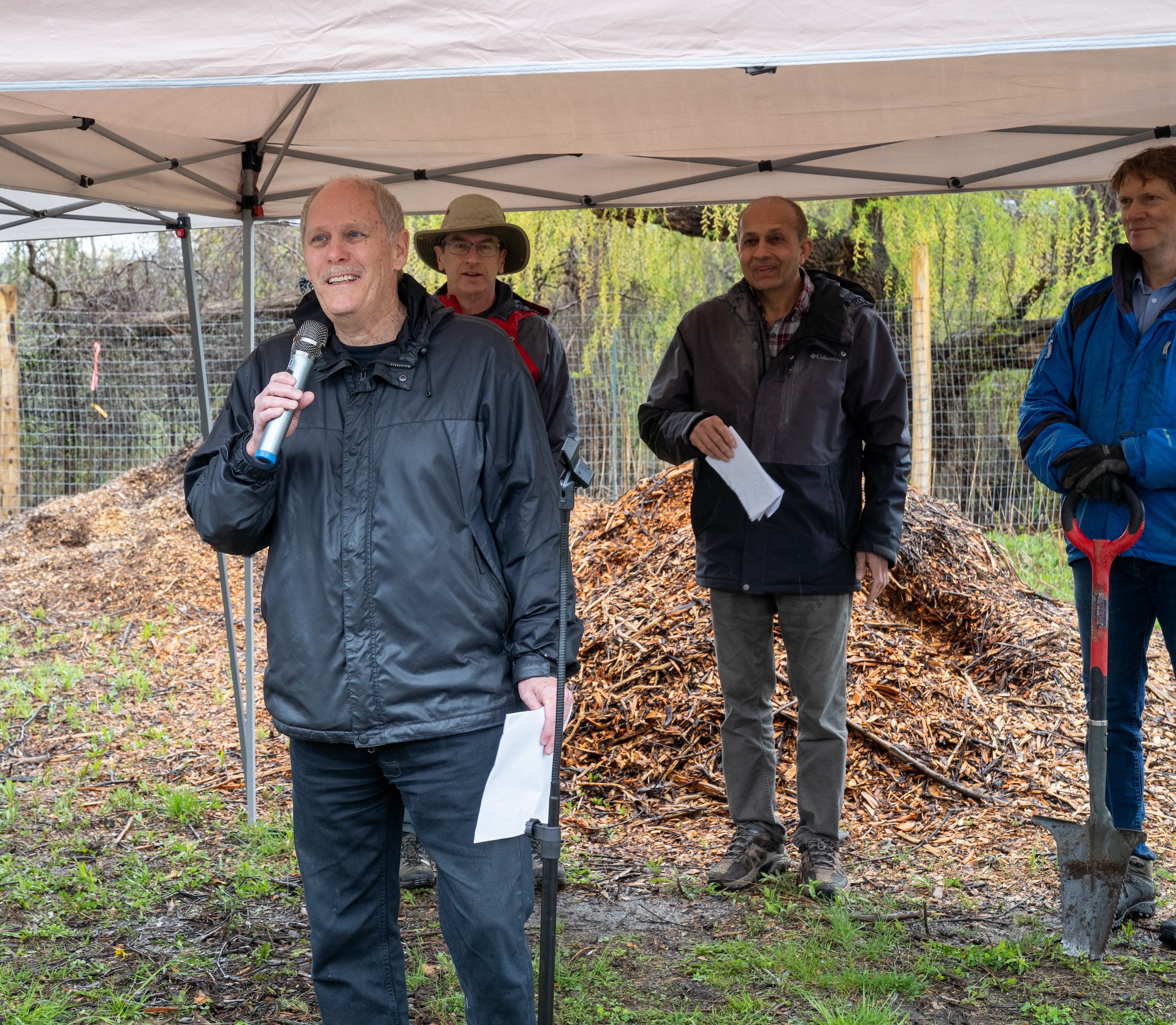 David Farrar, wearing a wet raincoat and standing under a temporary shelter, speaking into a mic, watched by researchers Bruce Newbold, Altaf Arain and Myles Sergeant.