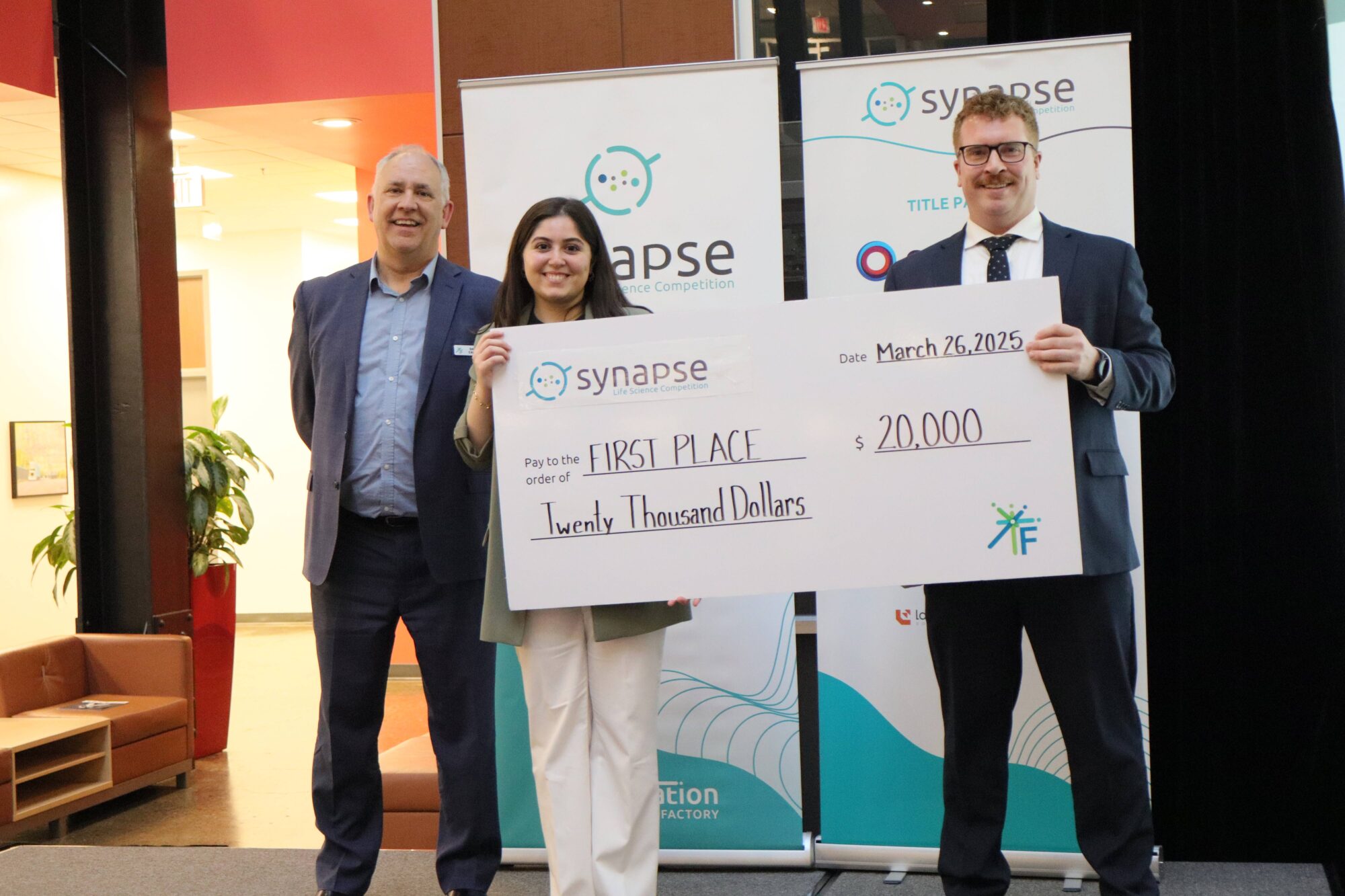 Two people in suits hold an oversized cheque that reads 'Synapse, First Place, Twenty Thousand Dollars.' A man in a suit stands behind them.