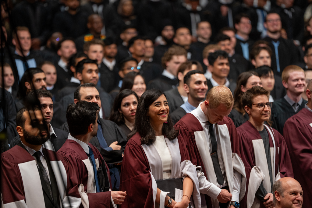Many people stand, all wearing graduation robes, facing the same direction. 