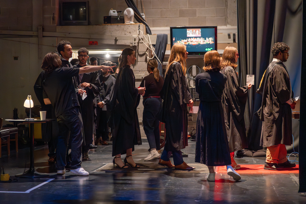 Backstage, several people in graduation robes stand in a short line. 