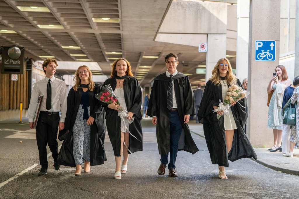 Five students in graduation robes walk together in a parking garage.