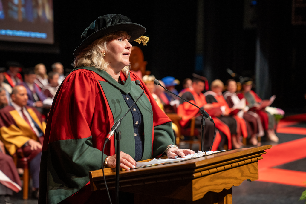 A woman in ceremonial robes stands at a podium, speaking. Many people are visible seated behind her on stage. 