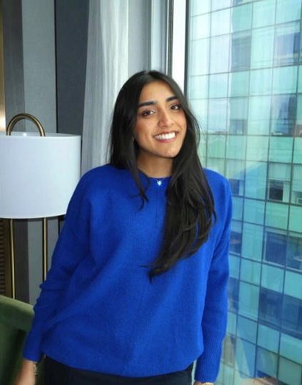 A young woman in a blue sweater smiles for the camera. A lamp and a glass window are behind her. 