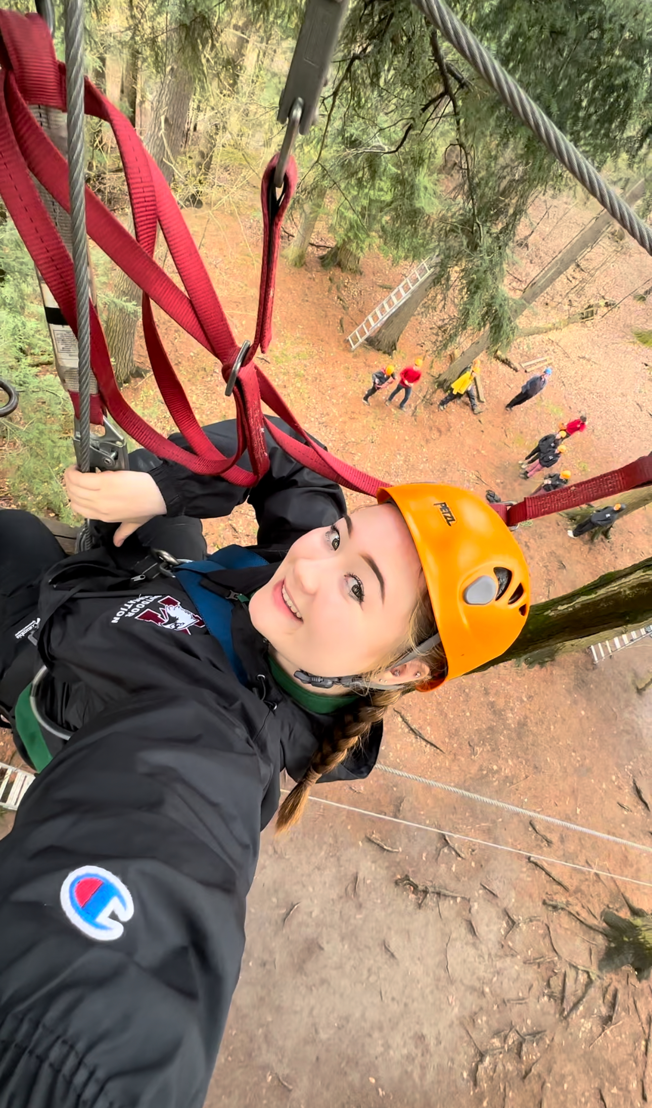 A woman in a harness and safety hat dangles above the ground, taking a selfie. People are visible standing on the ground behind and below her.