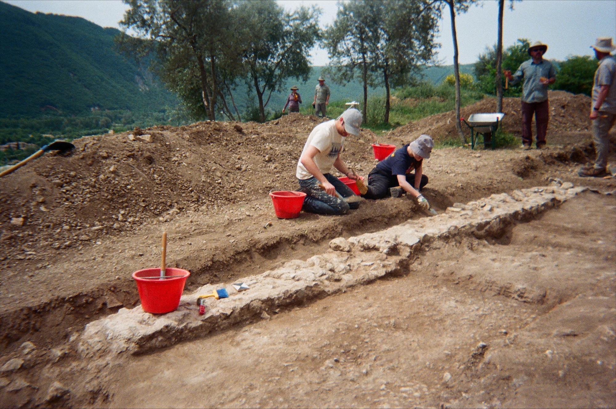 At an archaeological dig site, two people kneel in the dirt, focused on the trench in front of them. Several more people are standing in the background, and orange buckets and a shovel are in the dirt nearby. Behind all of the people is a green mountain range.