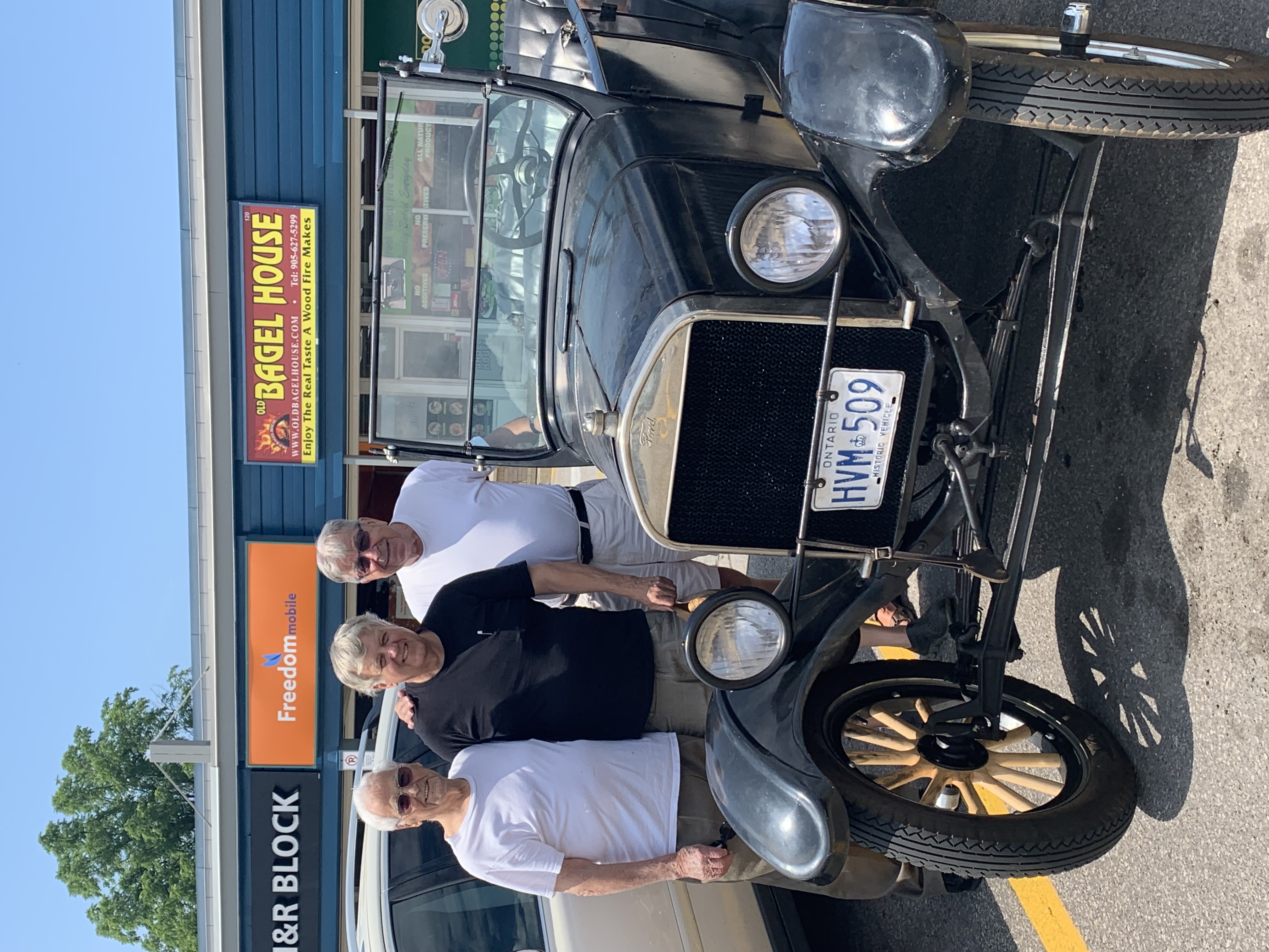 Three white-haired men stand beside a classic car in the parking lot in front of the Bagel House.