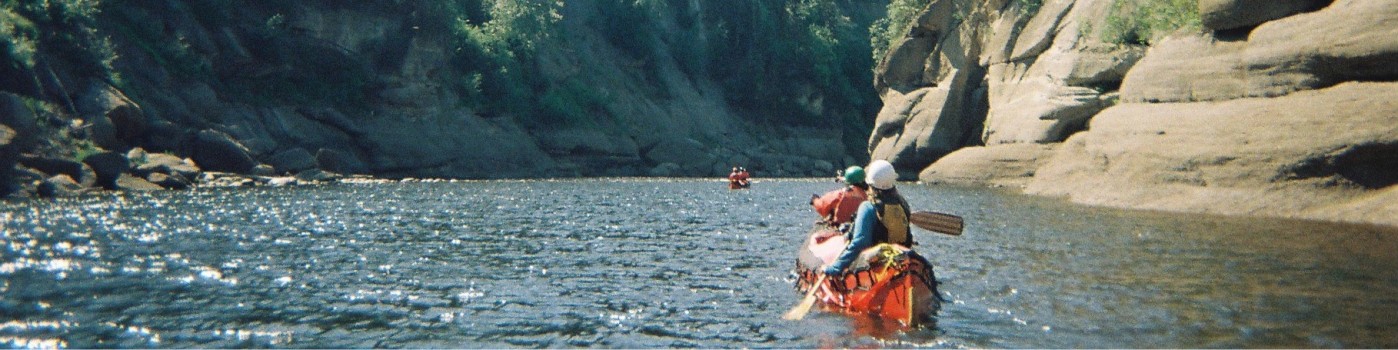 Two paddlers go down a river lined by tall rocks.