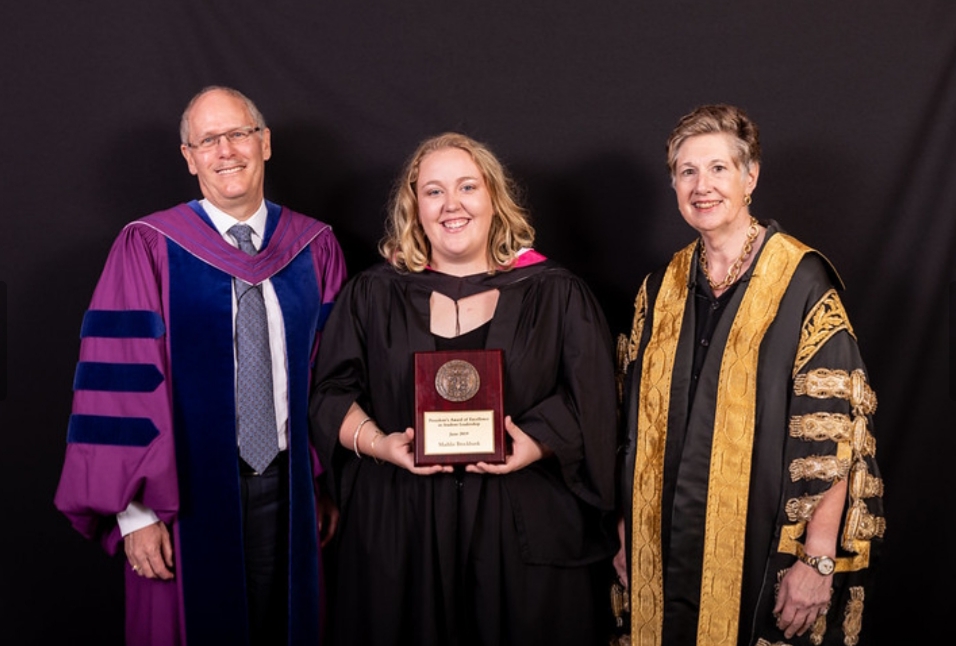 A woman in graduation robes holds a plaque, standing between two people in ceremonial robes.