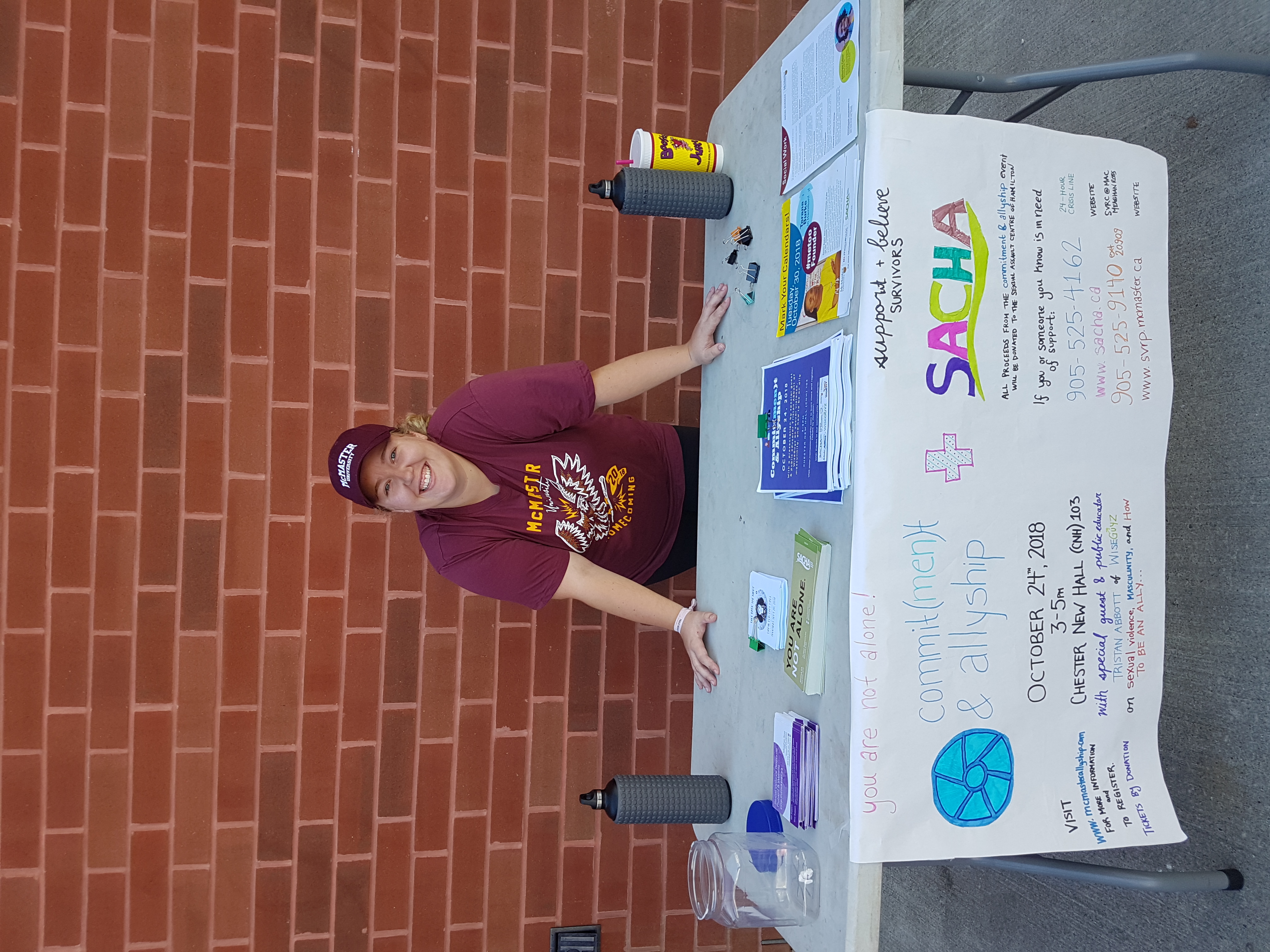 A woman stands outside at a folding table, set up with posters and water bottles.