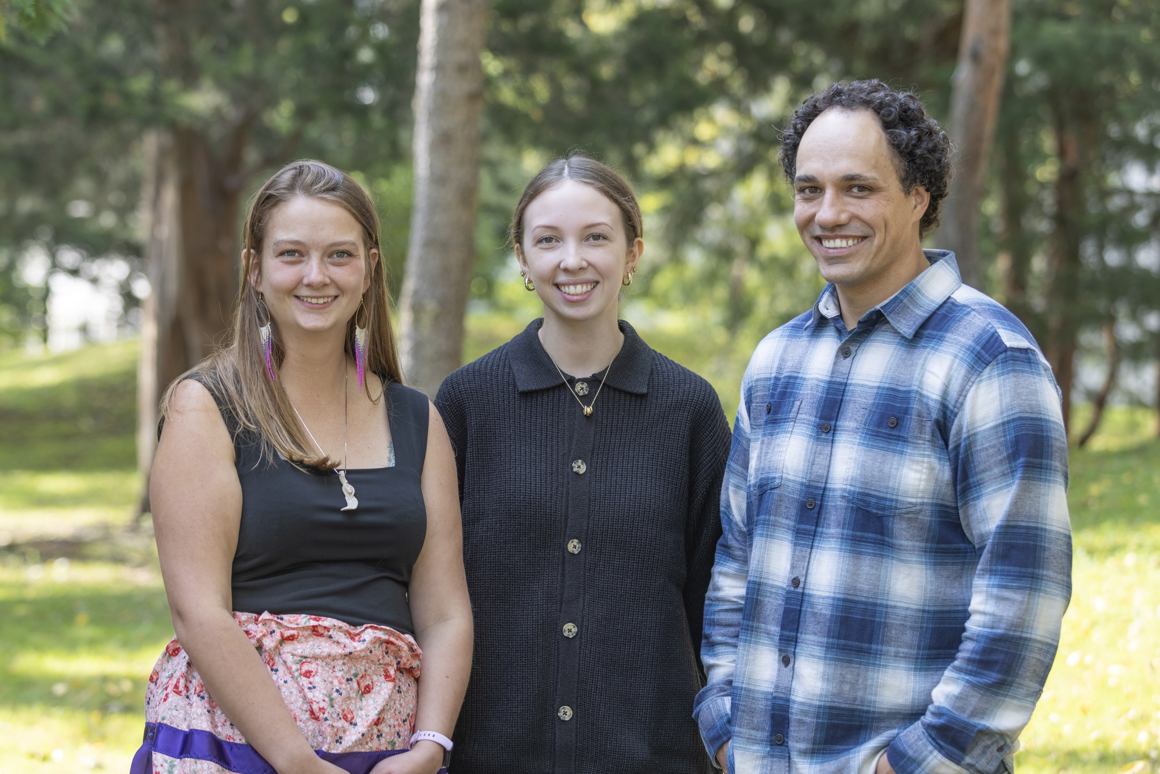 Two young women and a man stand together outside, smiling for the camera.
