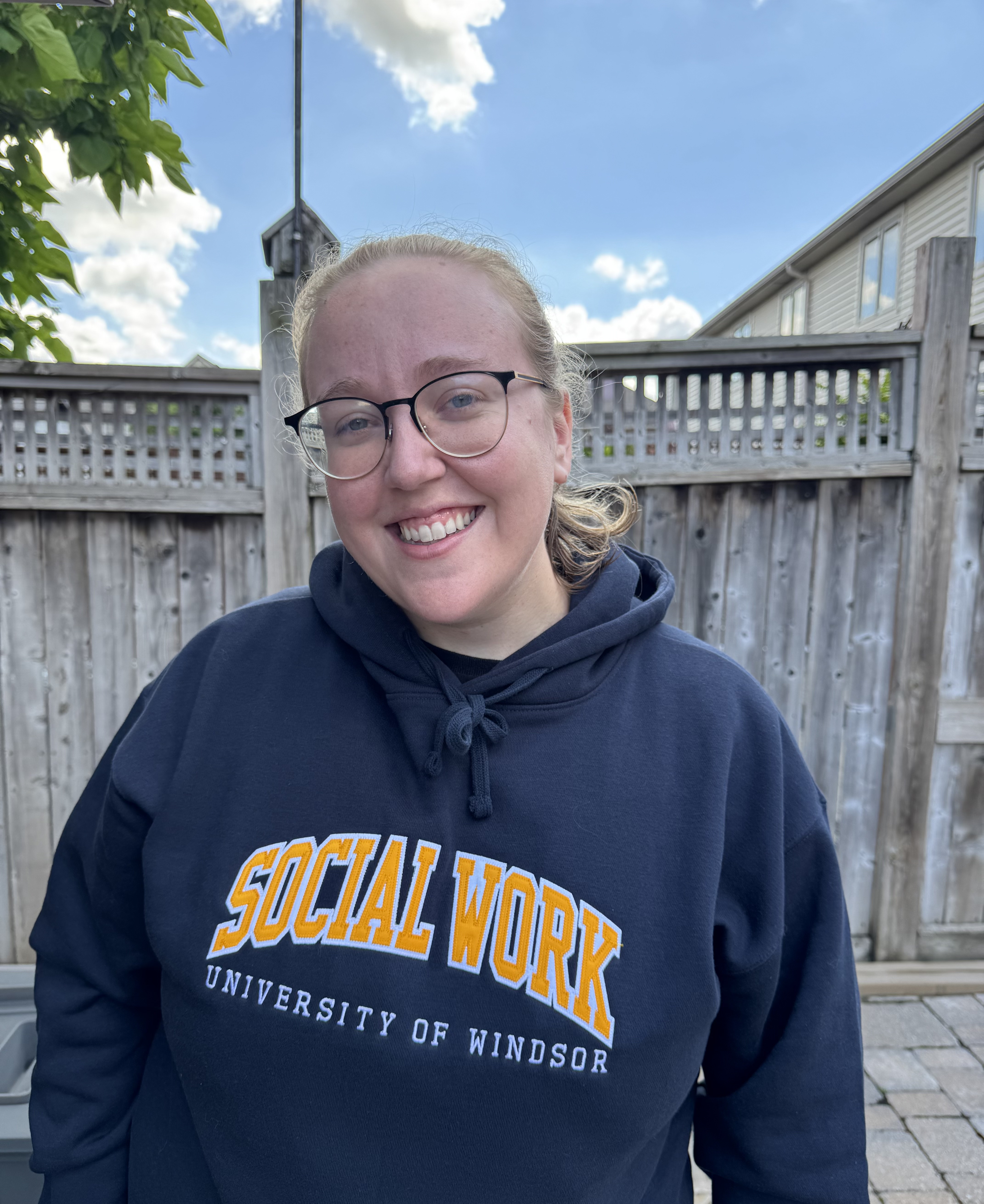 A woman stands on a deck outdoors, wearing a sweatshirt that reads 'Social Work, University of Windsor.'