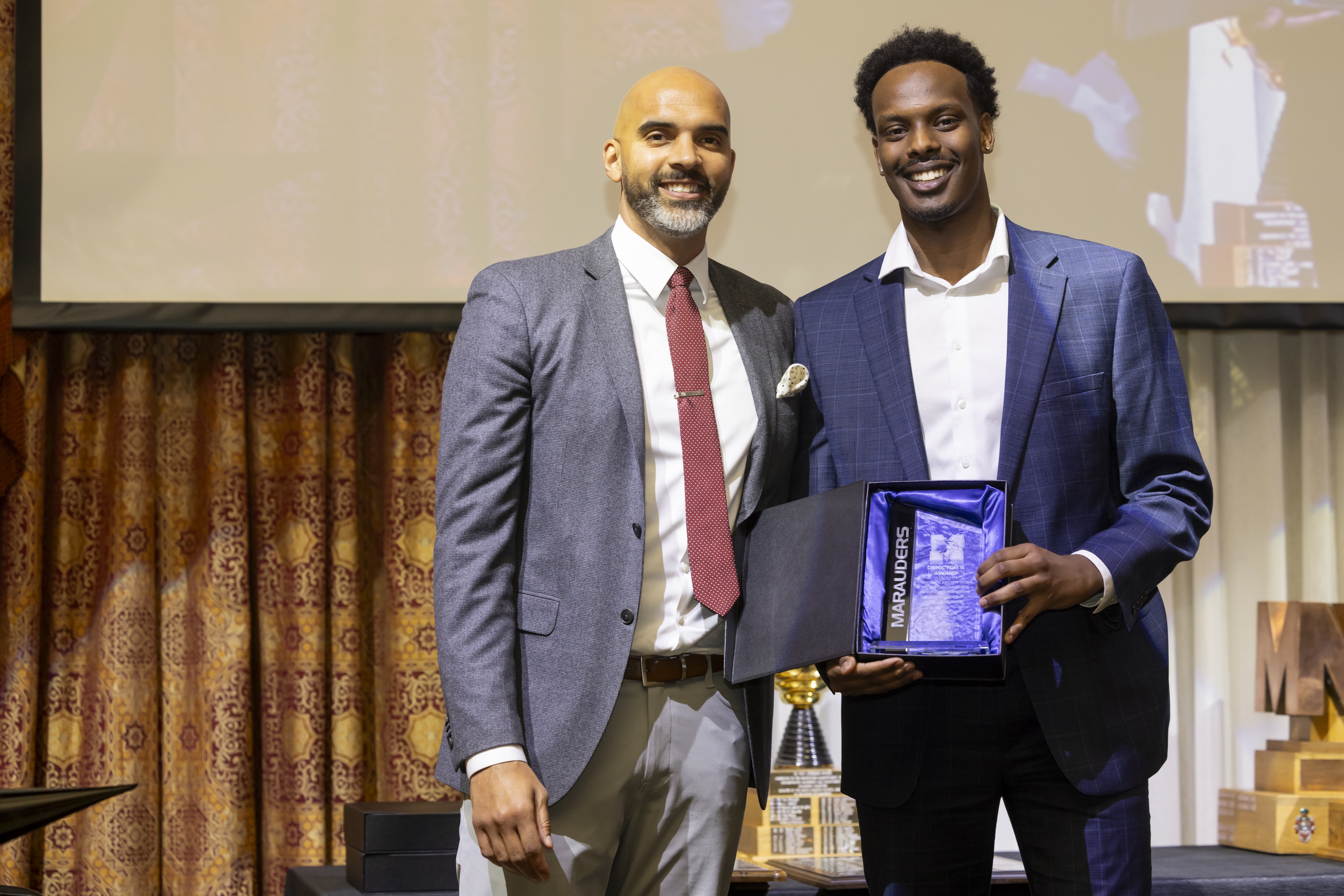 Two men stand on a stage in suits, one holding up an award. 