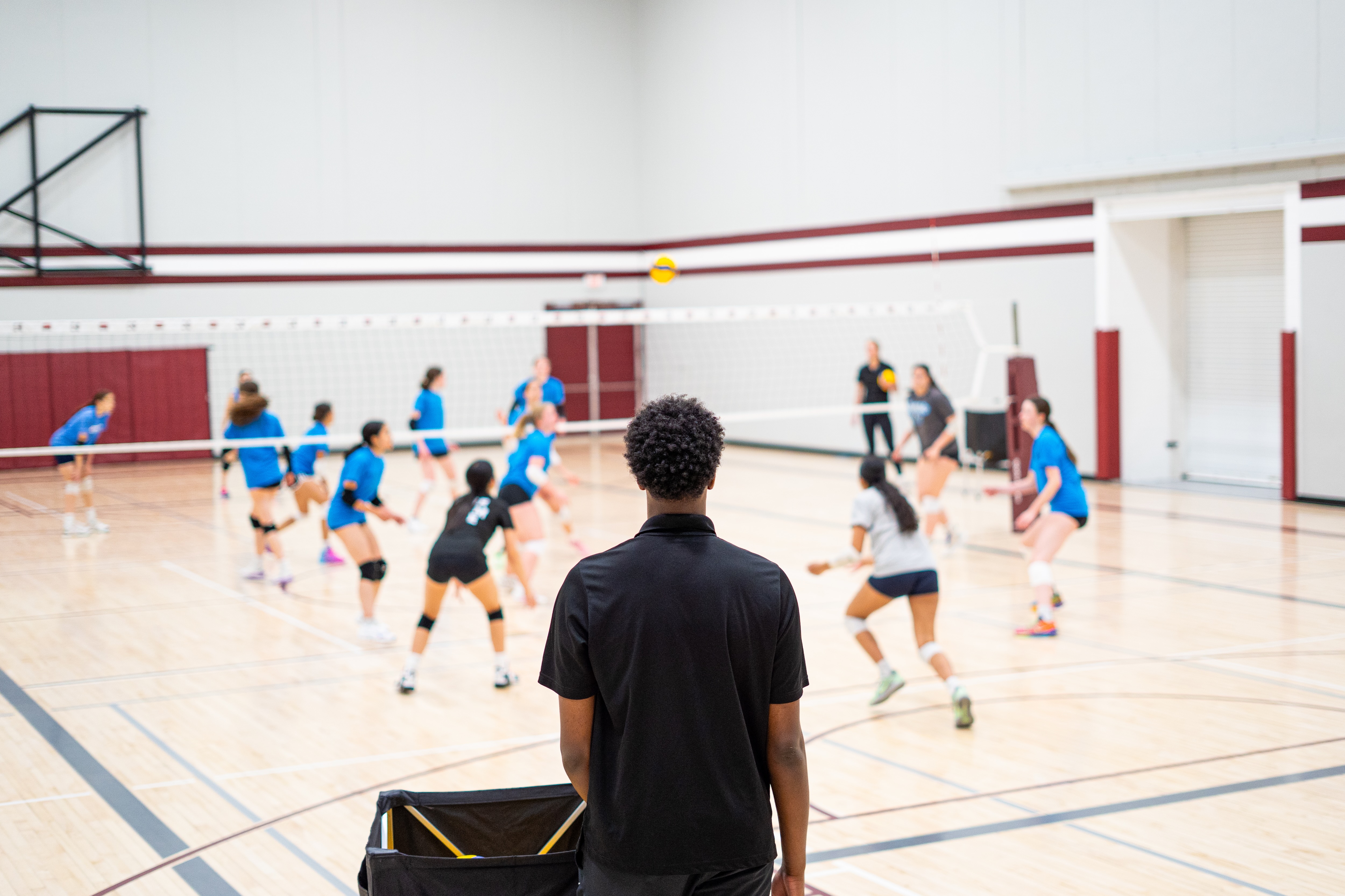A man stands on the edge of a volleyball court, watching a game.