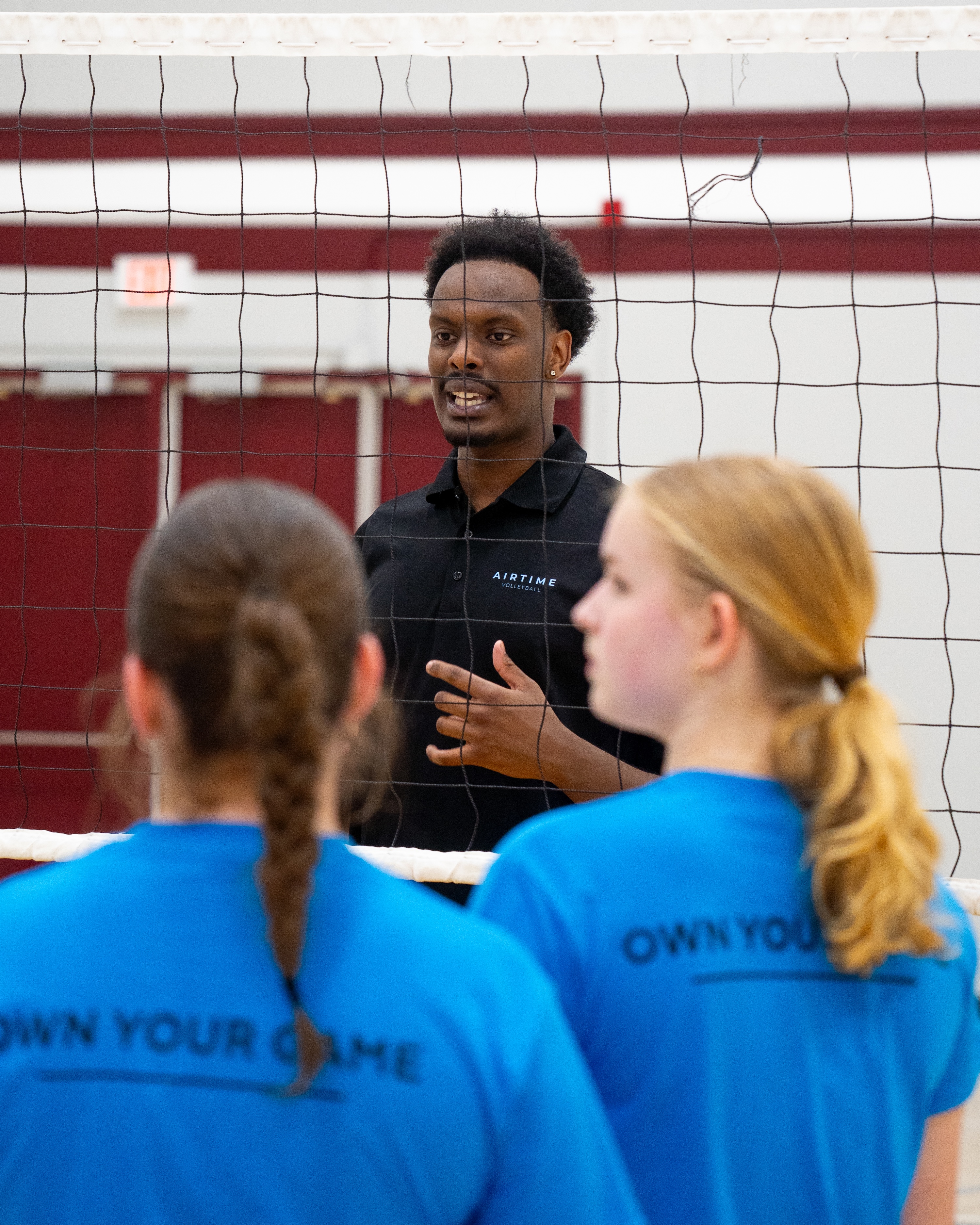 A man stands behind a volleyball net, speaking to volleyball players on the other side.