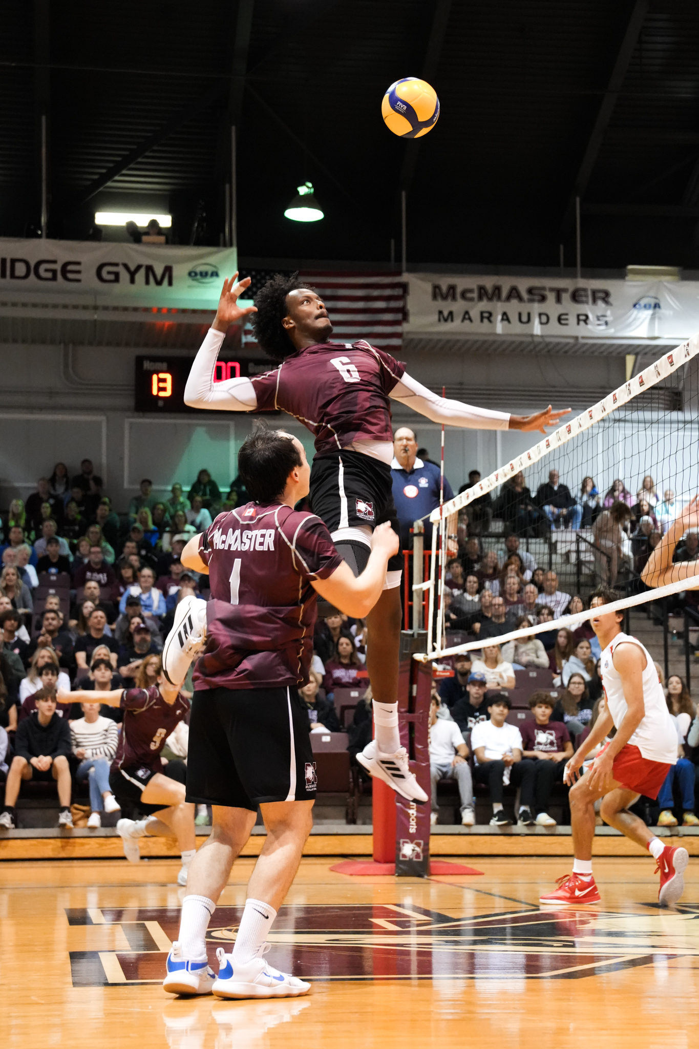 A man jumps to spike a volleyball during a volleyball game. Many spectators are visible in the back, sitting on bleachers.