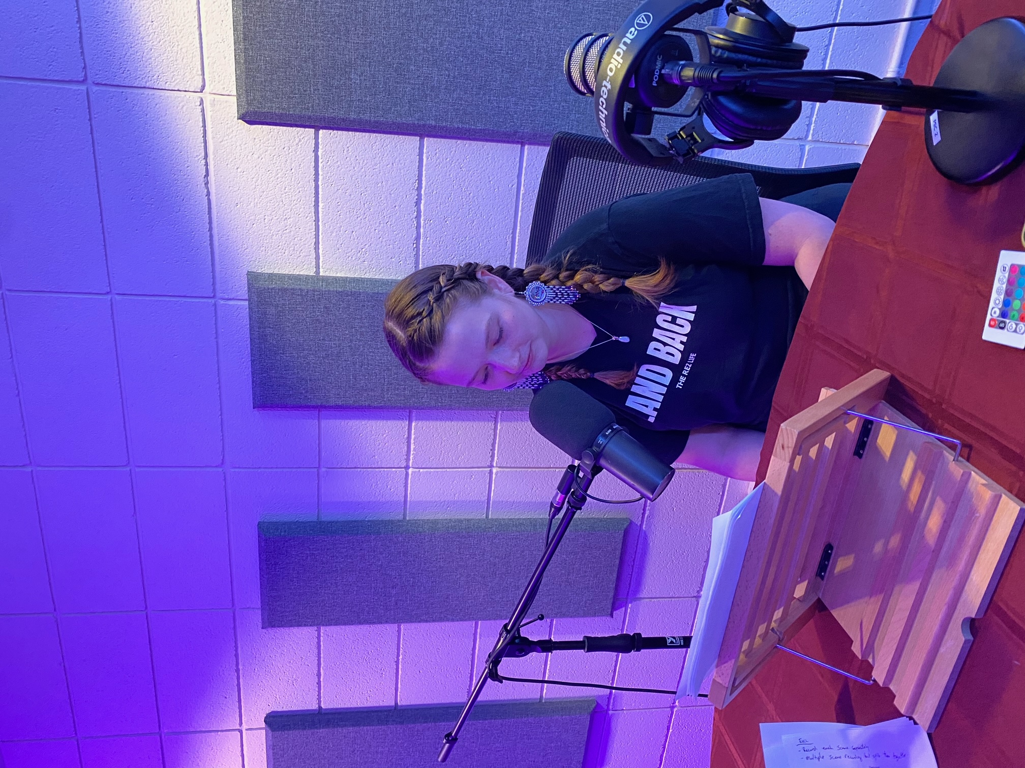 A woman sits in a recording booth, leaning in to a microphone.