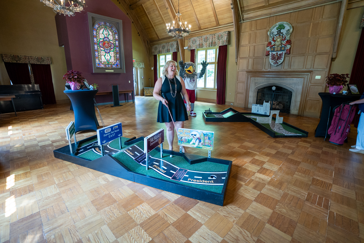 Susan Tighe laughs, holding a putter and standing at an indoor mini golf course in Alumni Memorial Hall as Mac the Marauder mascot holds up their arms behind her.
