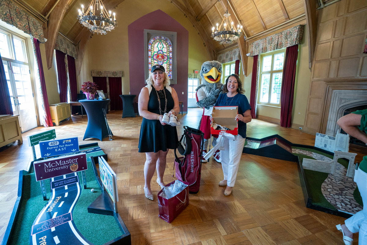 Susan Tighe holding a pair of new sneakers at the mini golf course set up in Alumni Memorial Hall, standing beside a staff member and Mac the Marauder mascot, with a golf bag beside her.